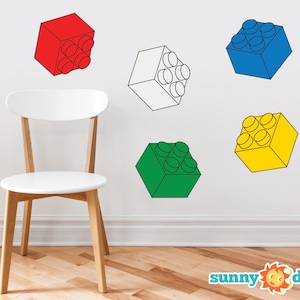 May include: Five colorful wall decals shaped like Lego blocks. The blocks are red, white, blue, green, and yellow. The decals are on a white wall with a white chair in front of them.