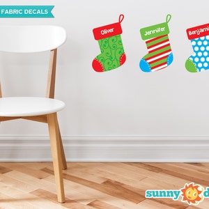 May include: Three reusable fabric Christmas stocking decals with names "Oliver", "Jennifer", and "Benjamin" in red lettering. The stockings are green, red and white striped, and blue with white polka dots.