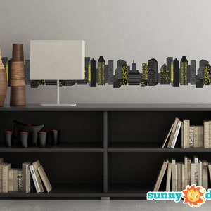May include: A black bookshelf with two shelves, filled with books. Two brown vases and a white lamp are on the top shelf. A black cityscape wall decal is on the wall behind the bookshelf. The decal features yellow windows and the text "sunny decals".