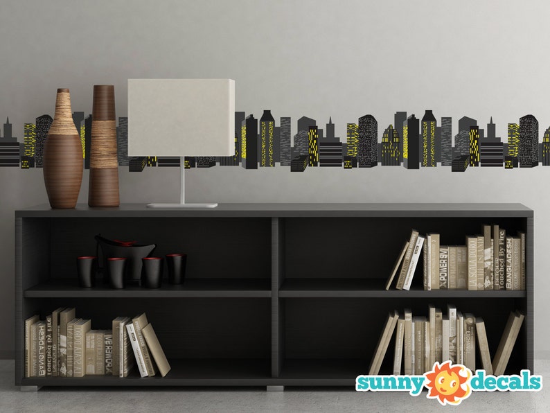 May include: A black bookshelf with two shelves, filled with books. Two brown vases and a white lamp are on the top shelf. A black cityscape wall decal is on the wall behind the bookshelf. The decal features yellow windows and the text "sunny decals".