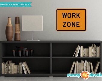 May include: A black bookshelf with two shelves, filled with books. On the top shelf, there are two vases, one brown and one beige. On the bottom shelf, there are more books and a black and red bowl. A white lamp sits on top of the bookshelf. A bright orange sign with black text reads "WORK ZONE" on a gray wall behind the bookshelf.