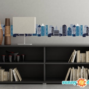 May include: A black bookshelf with two shelves, filled with books. Two vases with a wood grain pattern are on the top shelf, along with a white lamp. A cityscape wall decal with blue and gray buildings is on the wall behind the bookshelf. The text "sunny decals" is on the bottom of the image.