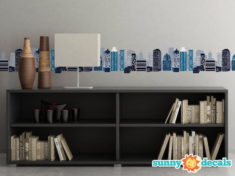 May include: A black bookshelf with two shelves, filled with books. Two vases with a wood grain pattern are on the top shelf, along with a white lamp. A cityscape wall decal with blue and gray buildings is on the wall behind the bookshelf. The text "sunny decals" is on the bottom of the image.