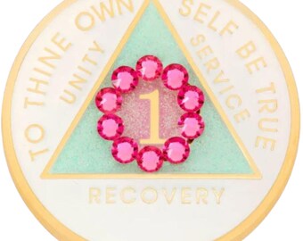 May include: A gold-toned round coin with a white background and a green triangle in the center. The triangle has the words "To Thine Own Self Be True" written around it. Inside the triangle is a pink circle of rhinestones surrounding the number "1" and the word "Recovery".