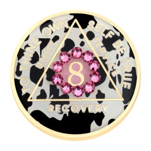 May include: A gold-rimmed medallion with a black and white cow print background. A white triangle with the words "SELF, SERVICE, BE TRUE" surrounds a pink number 8 encircled by pink gemstones. The words "TO THINE OWN UNITY" and "RECOVERY" are also present.