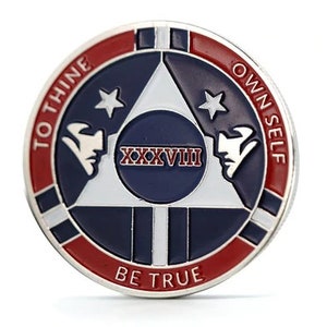 Op de afbeelding: Een zilverkleurige munt met een rood, wit en blauw ontwerp. De munt heeft een witte driehoek met twee sterren en de tekst "XXXVIII" in het midden. De tekst "To Thine Own Self Be True" staat in een cirkel rond de driehoek.
