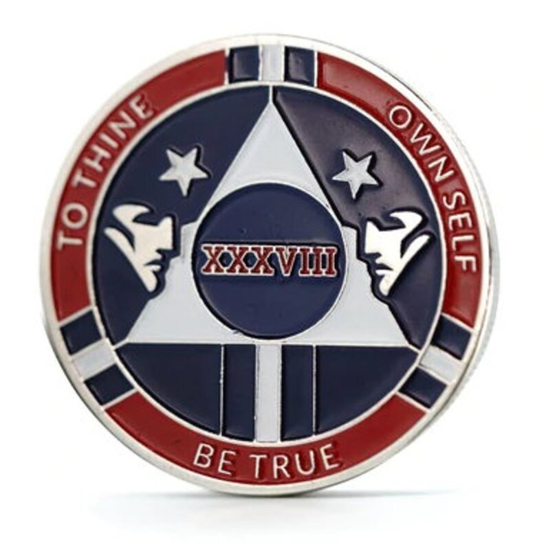Op de afbeelding: Een zilverkleurige munt met een rood, wit en blauw ontwerp. De munt heeft een witte driehoek met twee sterren en de tekst "XXXVIII" in het midden. De tekst "To Thine Own Self Be True" staat in een cirkel rond de driehoek.