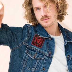 May include: A dark blue denim jacket with a black and red patch. The patch displays the letters "NA" within a circle, accompanied by the phrases "NEVER ALONE" and "NEVER AGAIN". The jacket has a traditional design, complete with button fastenings and pockets.