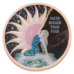Könnte beinhalten: Eine runde goldfarbene Münze mit schwarzem Hintergrund. Die Münze zeigt eine Frau, die zu einer Sonne mit rosafarbenem Zentrum greift. Die Sonne ist von weißen Strahlen und einem schwarzen Sternenhimmel umgeben. Der Text "FAITH BIGGER THAN FEAR" ist auf der rechten Seite der Münze aufgedruckt.