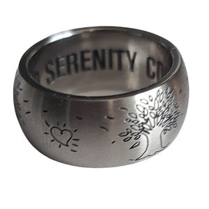 May include: A silver ring with the words "Serenity Now" engraved on the inside. The outside of the ring features a tree, a heart, and a sun.