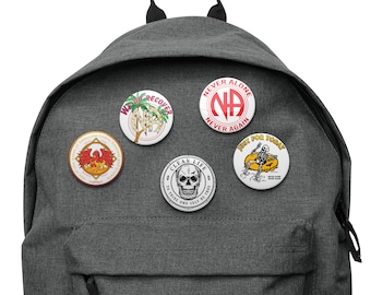 Puede incluir: Una mochila gris con cinco pines redondos unidos a ella. Los pines presentan varios diseños, incluyendo un fénix, una calavera, un esqueleto y las letras "NA" en un círculo. El texto en los pines dice "Never Alone, Never Again", "Just for Today", "Clean Life", "To Thine Own Self Be True", y "We Do Recover".