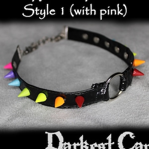 Puede incluir: Dos gargantillas de cuero negro con anillos de metal plateado y pinchos de colores. Las gargantillas están etiquetadas como "Rainbow Pride Style 1 (con rosa)" y "Rainbow Pride Style 2 (con rosa)".