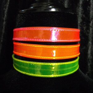 May include: Three colorful chokers in neon orange, green, and pink. The chokers are made of a shiny material and are stacked on top of each other. The text "Darkest Candy Designs" is at the bottom.