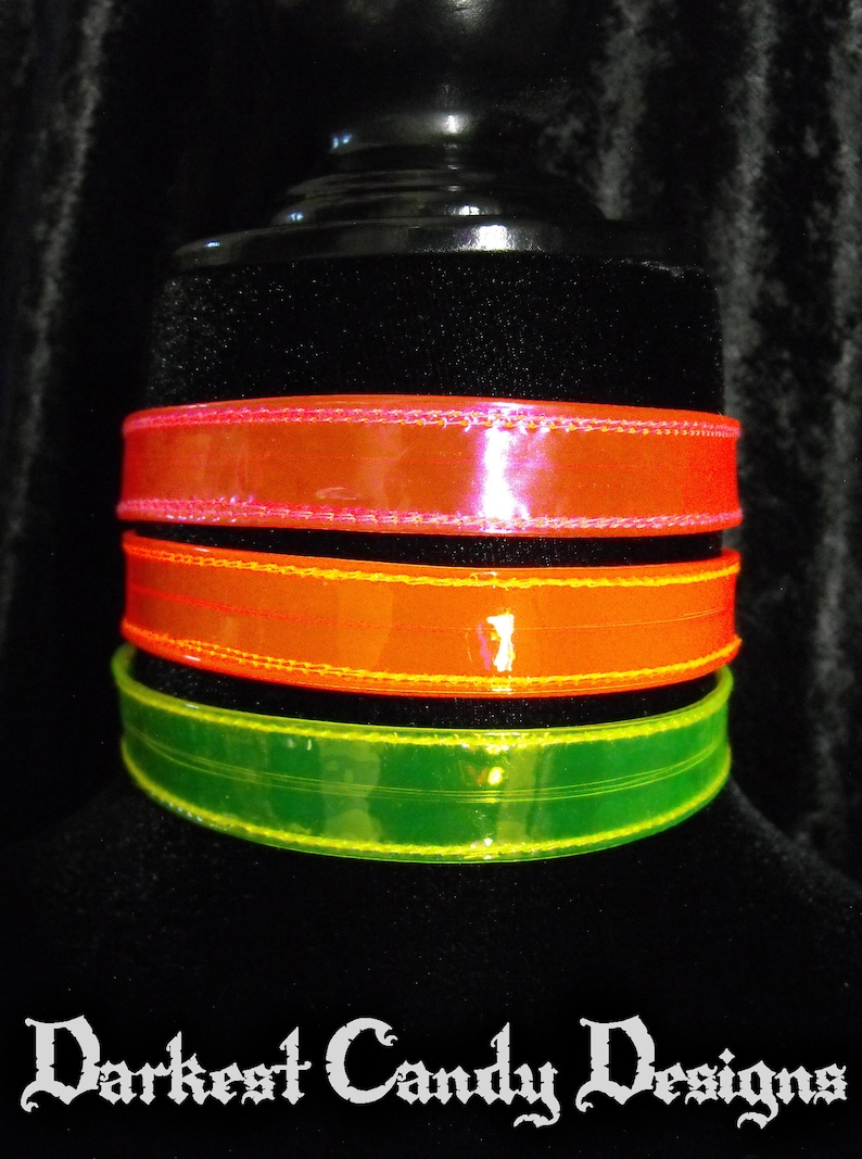 May include: Three colorful chokers in neon orange, green, and pink. The chokers are made of a shiny material and are stacked on top of each other. The text "Darkest Candy Designs" is at the bottom.