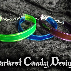 May include: Three choker necklaces in green, blue, and purple. Each choker has a shiny, iridescent band with silver-colored hardware and a chain clasp. The text "Darkest Candy Designs" is at the bottom of the image.
