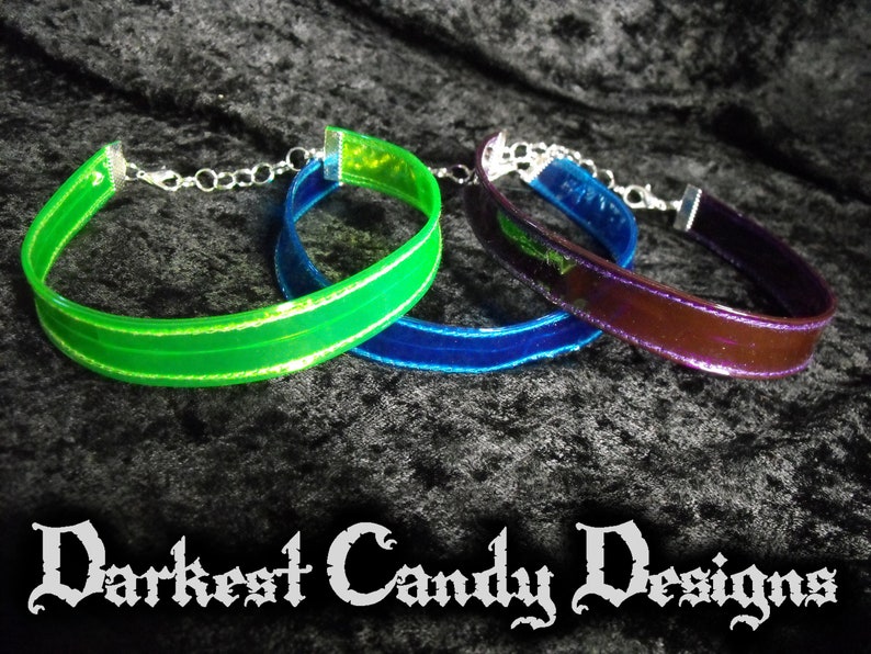 May include: Three choker necklaces in green, blue, and purple. Each choker has a shiny, iridescent band with silver-colored hardware and a chain clasp. The text "Darkest Candy Designs" is at the bottom of the image.