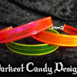 May include: Three colorful chokers in red, orange, and green, each with a shiny, iridescent finish. The chokers are made of a flexible material and have a silver clasp. The text "Darkest Candy Designs" is at the bottom.