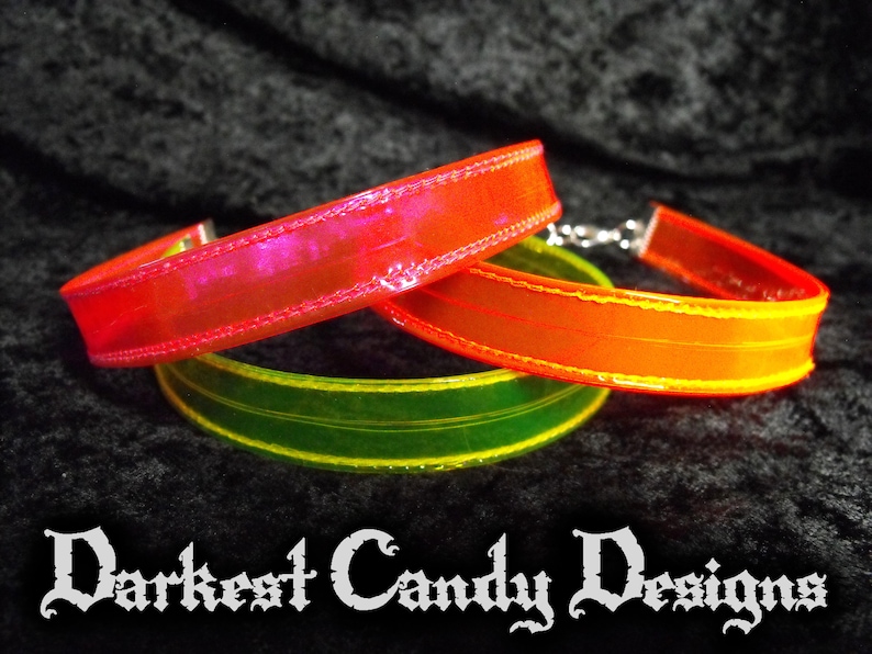 May include: Three colorful chokers in red, orange, and green, each with a shiny, iridescent finish. The chokers are made of a flexible material and have a silver clasp. The text "Darkest Candy Designs" is at the bottom.