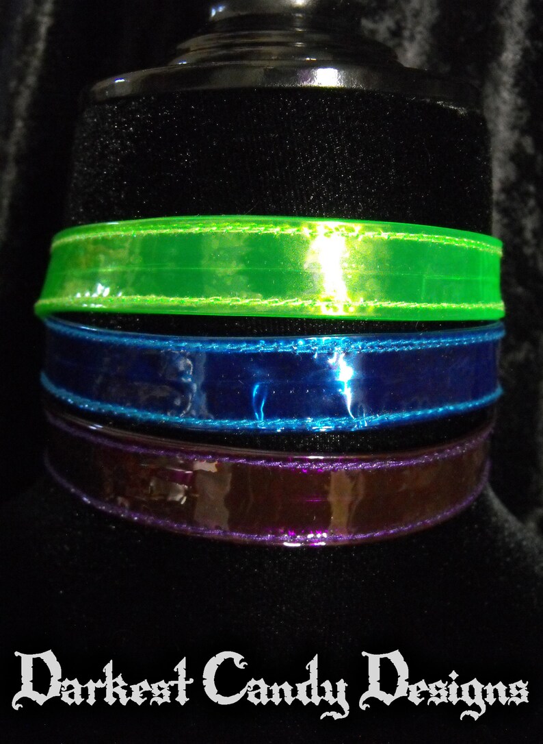 May include: Three colorful chokers in green, blue, and purple. Each choker is made of a shiny, transparent material with a stitched edge. The text "Darkest Candy Designs" is at the bottom.