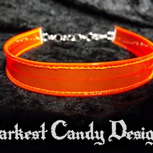 May include: Bright orange choker necklace with a silver chain clasp. The choker is made of a translucent material with a stitched border. The text "Darkest Candy Designs" is at the bottom of the image.