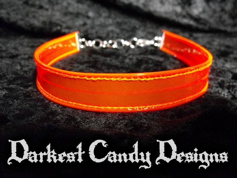 May include: Bright orange choker necklace with a silver chain clasp. The choker is made of a translucent material with a stitched border. The text "Darkest Candy Designs" is at the bottom of the image.