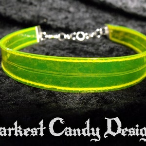 May include: A neon green choker necklace with yellow trim and silver-colored chain clasp. The choker has a transparent appearance and features three thin, parallel lines. The text "Darkest Candy Designs" is at the bottom.