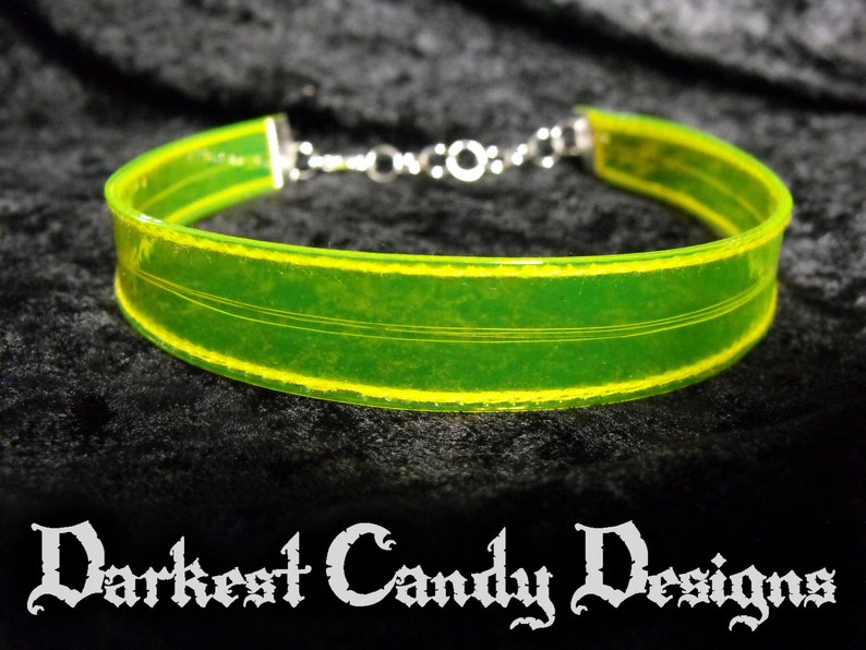 May include: A neon green choker necklace with yellow trim and silver-colored chain clasp. The choker has a transparent appearance and features three thin, parallel lines. The text "Darkest Candy Designs" is at the bottom.