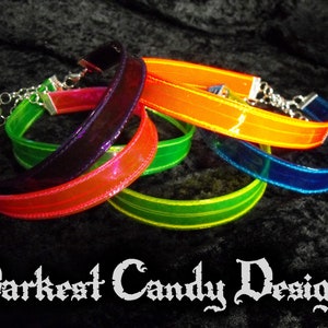 May include: A collection of colorful chokers in shades of red, orange, green, and blue. Each choker features a clear, iridescent band with a silver chain clasp. The text "Darkest Candy Designs" is displayed at the bottom.