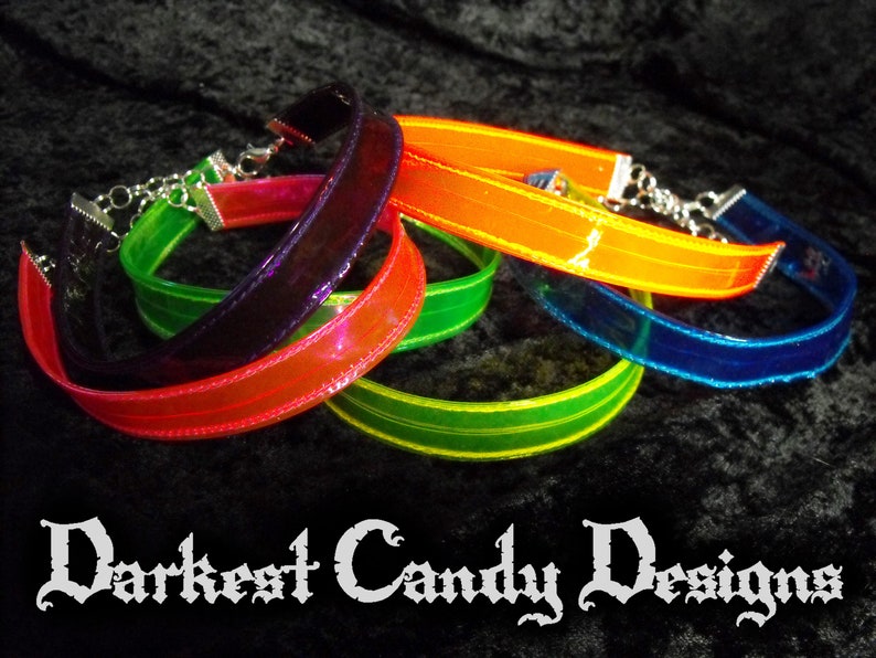 May include: A collection of colorful chokers in shades of red, orange, green, and blue. Each choker features a clear, iridescent band with a silver chain clasp. The text "Darkest Candy Designs" is displayed at the bottom.