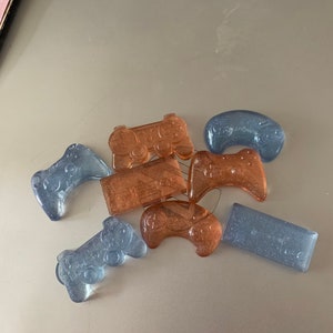 May include: A collection of seven small, translucent, glitter-filled game controllers in blue and brown shades. The controllers are shaped like various gaming consoles.