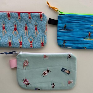 May include: Three rectangular zippered pouches with unique patterns. One features swimmers in red swimsuits doing yoga poses on a blue background. Another has paddleboarders on a blue water background. The third has swimmers on a light green background.