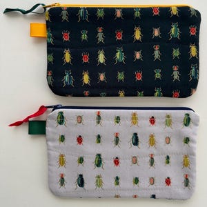 May include: Two rectangular zippered pouches. One is navy blue with a pattern of colorful beetles, a yellow zipper, and a green pull tab. The other is light gray with a similar beetle pattern, a blue zipper, and a red pull tab.