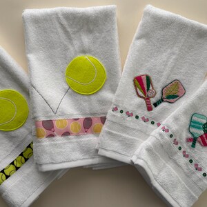 May include: Four white terry cloth towels featuring sports themes. Two towels display yellow tennis ball appliqués and patterned trim. Two towels feature embroidered pickleball paddles and balls with decorative ribbon borders.
