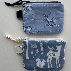 May include: Two small, square coin purses. The top purse is light blue with a deer pattern. The bottom purse is light blue with a deer, squirrel, and floral pattern. Both purses have zippers and small loops.