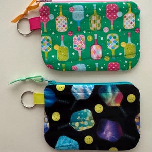 May include: Two small, rectangular zippered coin purses. One is green with pickleball paddle designs in various colors. The other is black with pickleball paddle designs and yellow pickleballs. Each has a key ring.