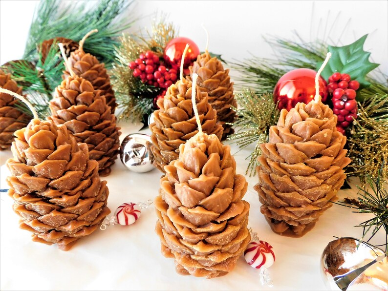 May include: Five brown pine cone shaped candles with white wicks. The candles are arranged on a white surface with green pine branches and red and silver ornaments.