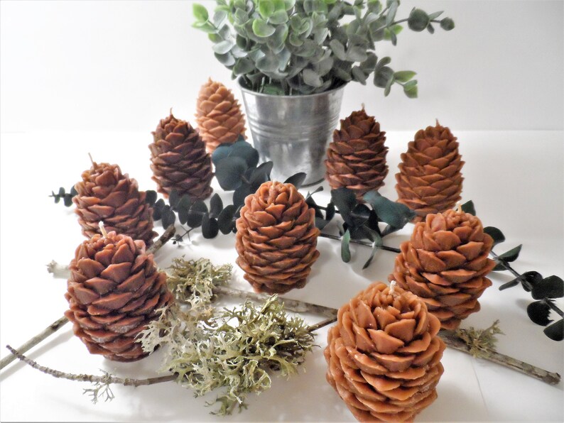 May include: A group of eight brown pine cone shaped candles on a white surface with green eucalyptus leaves and brown moss.