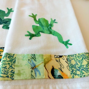 May include: Two white tea towels with green frog silhouettes and a patchwork border of green, yellow, and blue fabric with a butterfly, dragonfly, and bird designs.