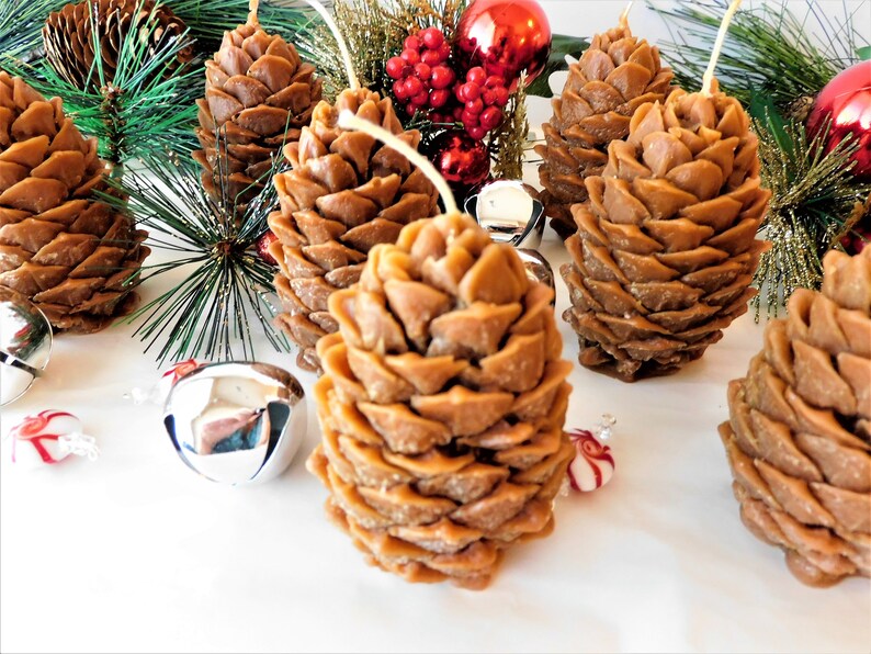May include: A set of six brown pine cone shaped candles. The candles are arranged on a white surface with silver bells and red and green holiday decorations.