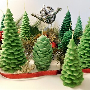May include: A collection of green pine tree shaped candles, some with a pine cone design, arranged on a white surface with red ribbon and gold and green holiday decorations.