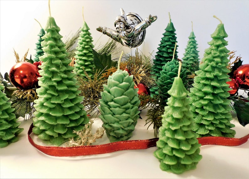 May include: A collection of green pine tree shaped candles, some with a pine cone design, arranged on a white surface with red ribbon and gold and green holiday decorations.