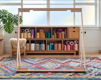 Op de afbeelding: Een groot houten weefgetouwframe in een lichte kleur met een rechthoekige structuur en inkepingen aan de boven- en onderbalk. Het frame staat voor een houten opbergmeubel gevuld met diverse klossen gekleurd garen.