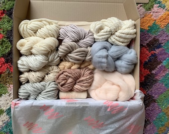 May include: A box filled with various shades of yarn and wool, including white, cream, light pink, light brown, and gray. The yarn is arranged in a neat and organized manner, ready for crafting projects.