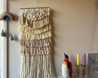 May include: A cream-colored, layered macrame wall hanging with a textured design. The wall decor features a variety of textures and lengths, with a row of thick, knotted fringe at the bottom. A decorative penguin figurine and candles are on a nearby shelf.