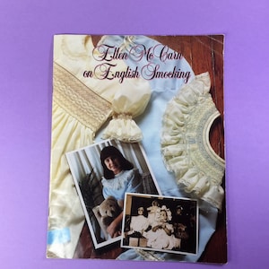 May include: A book titled "Ellen Mc Carn on English Smocking" with a yellow and white smocked dress on the cover. The book also features a photo of a young girl wearing a blue smocked dress and a teddy bear.