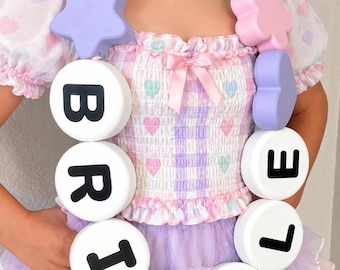 May include: A chunky necklace featuring large, matte-finish beads in pastel pink, purple, and mint green, shaped like hearts and stars. The necklace includes white circular beads with the black letters spelling out BRIELLE.