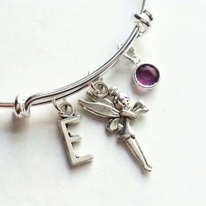 Könnte beinhalten: Silberarmband mit einem lilafarbenen Edelstein, einem Feen-Anhänger und einem Buchstaben E-Anhänger.