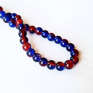 May include: A strand of round beads in a mix of red and blue. The beads are arranged in a loop against a white background. The beads appear to be made of glass or a similar material.