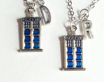 May include: Two silver-toned necklaces with blue and silver Tardis pendants. Each pendant has the words "POLICE BOX" on top. One necklace has a "D" charm, the other has an "R" charm. The necklaces are on a white background.