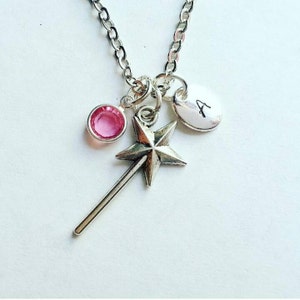 May include: Silver-toned necklace with a chain and multiple charms. Charms include a pink gemstone, a star-shaped wand, and a small oval pendant with the letter "A". The necklace is displayed on a white background.
